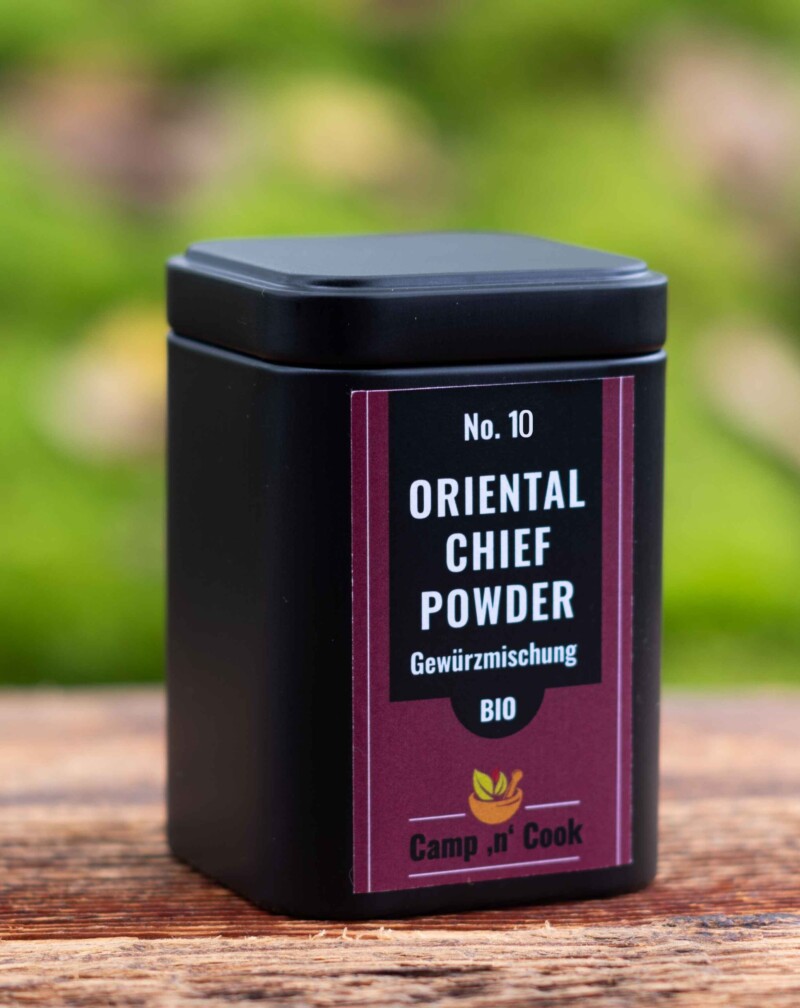 No. 10 Oriental Chief Powder bio Gewürzmischung steht auf dem auberginefarbenem Etikett. Es ist auf einer quadratischen schwarzen Dose angebracht, die auf einem Holzbrett steht. Der Hintergrund ist grün von Blättern.