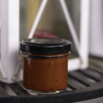Chilipaste Knock Out BIO - in einem kleinen Sturzglas mit schwarzem Deckel ist eine dunkelrote Paste zu erkennen. Das Glas steht auf einem Grillrost, im Hintergrund steht eine weiße Laterne.