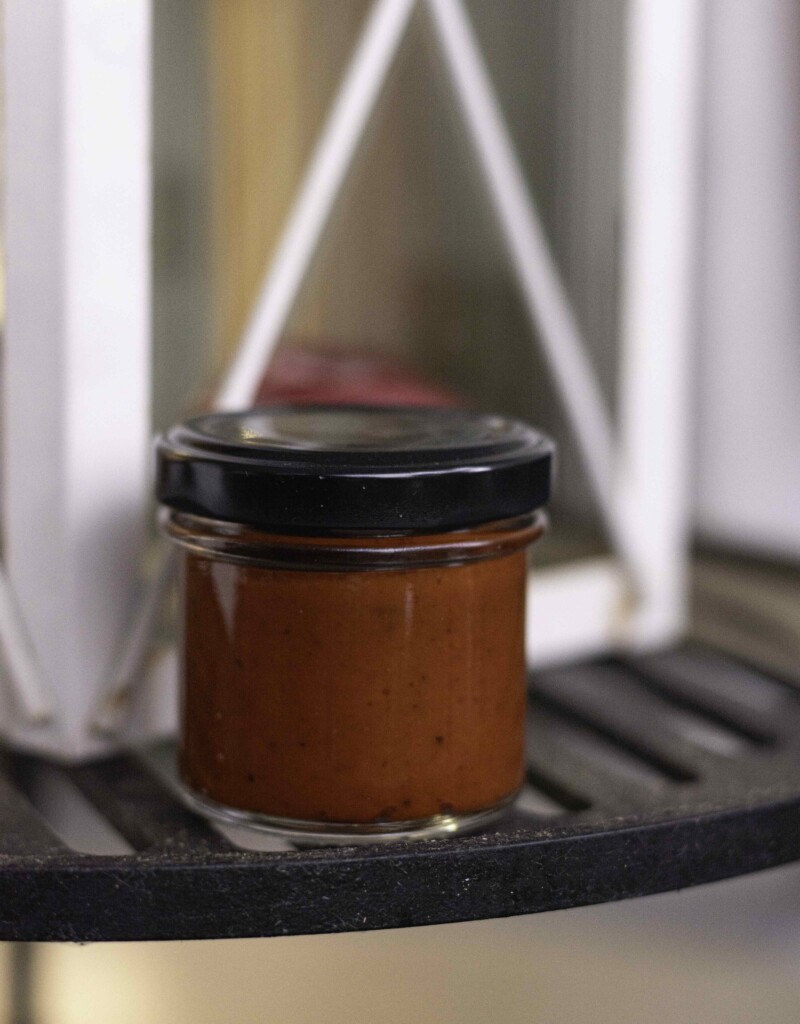 Chilipaste Knock Out BIO - in einem kleinen Sturzglas mit schwarzem Deckel ist eine dunkelrote Paste zu erkennen. Das Glas steht auf einem Grillrost, im Hintergrund steht eine weiße Laterne.
