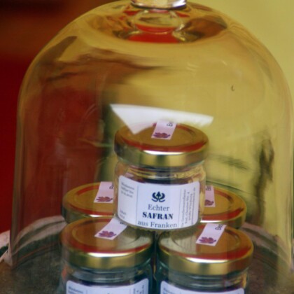 Safran aus Franken. 5 kleine Gläschen mit Safranfäden, versehen mit einem weißen Etikett und goldenem Deckel stehen unter einer Glasglocke.